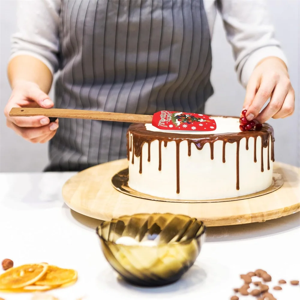 A hand is using a festive Christmas silicone spatula with a wooden handle to decorate a cake.
