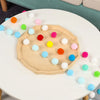 A colorful felt ball garland with various pastel colors is displayed on a white table.