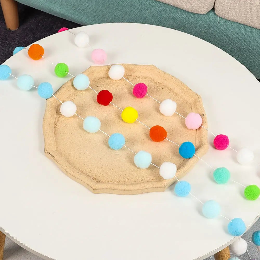 A colorful felt ball garland with various pastel colors is displayed on a white table.