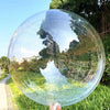 A clear PVC party balloon reflects trees and greenery in a natural outdoor setting.