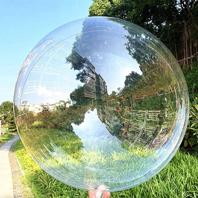 A clear PVC party balloon reflects trees and greenery in a natural outdoor setting.