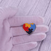 A hand wearing a light purple glove holds a heart-shaped puzzle ribbon brooch pin with rainbow colors.