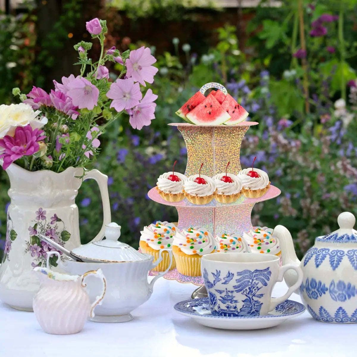 A 3-tier cardboard cupcake stand decorated with pink and gold accents, set on a white table amidst greenery.