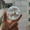 A hand holds a clear crystal ball water feature ornament against a blurred building background.