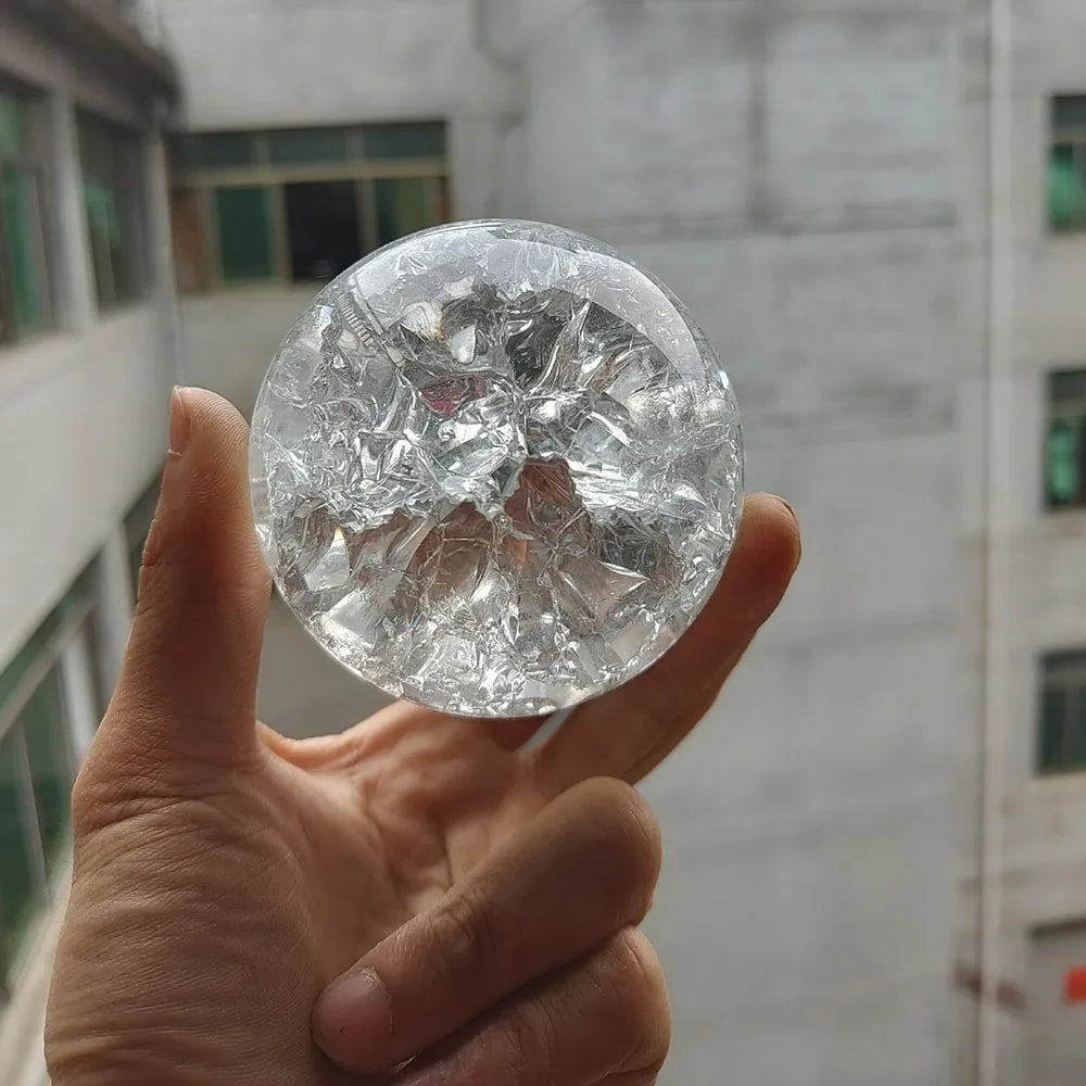 A hand holds a clear crystal ball water feature ornament against a blurred building background.