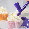 A purple pearl ball applicator tool is shown decorating a pink cupcake with white frosting.