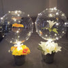 Two clear balloons filled with flowers, one yellow and one white, illuminated by string lights.