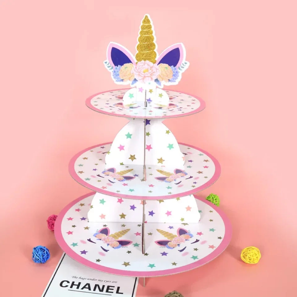 A three-tier pink and purple cardboard cupcake stand decorated with a gold unicorn topper and pastel floral accents.