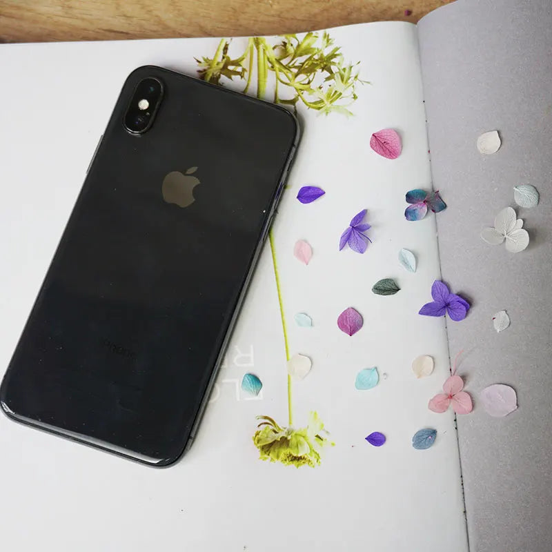 A black smartphone lies on a white page surrounded by scattered dried purple and green hydrangea petals.