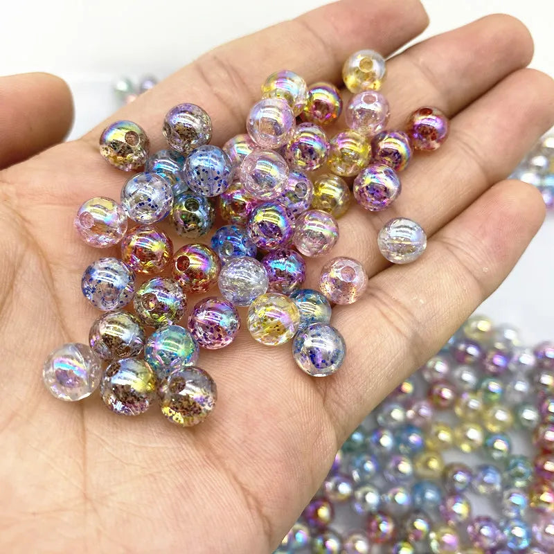 A hand holds a cluster of iridescent, round acrylic beads in various shades of purple, pink, and gold for jewelry making.