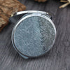 A silver mini double-sided folding makeup mirror with an engraved floral pattern resting on a dark wooden surface.