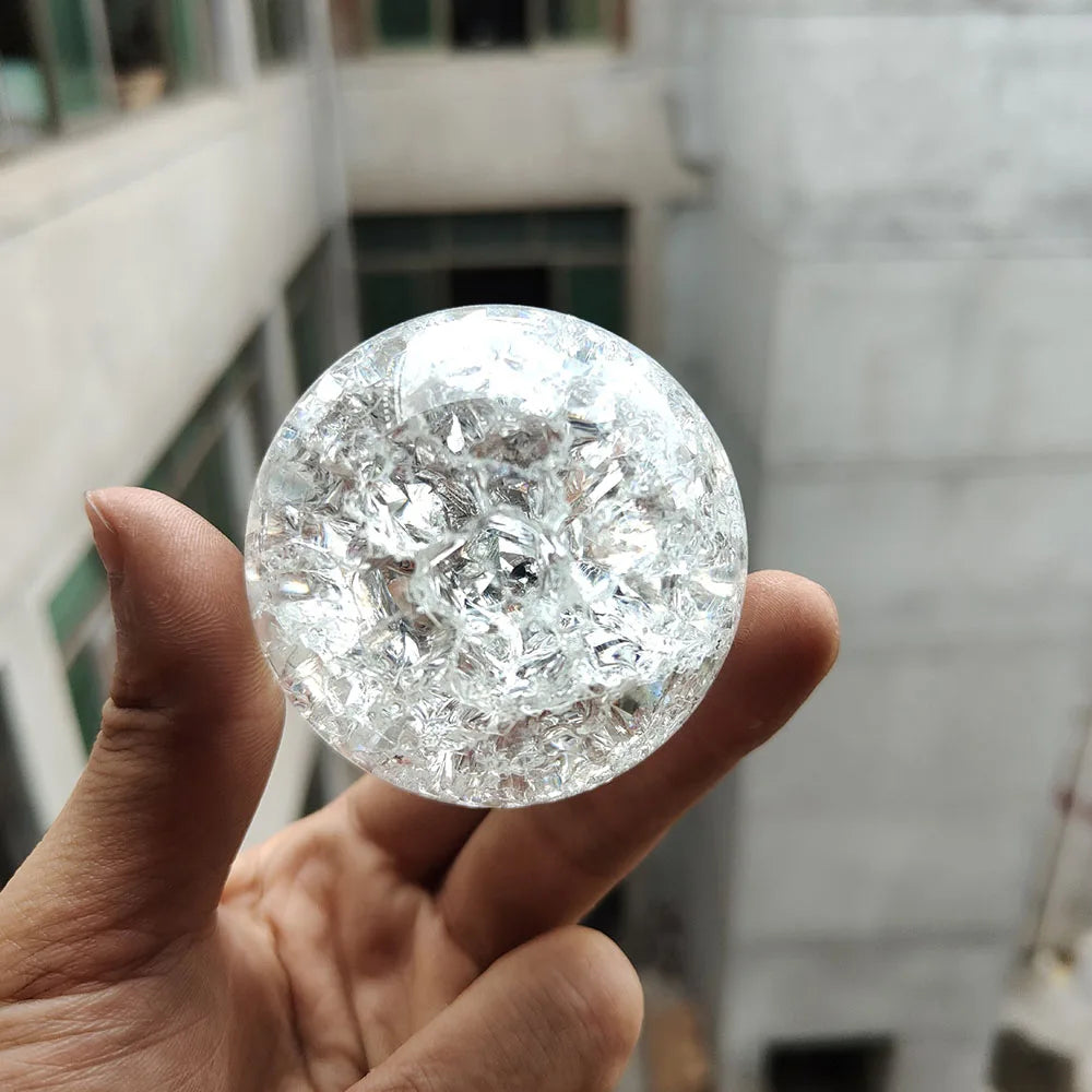 A hand holds a clear, spherical crystal ball water feature ornament against a blurred architectural background.