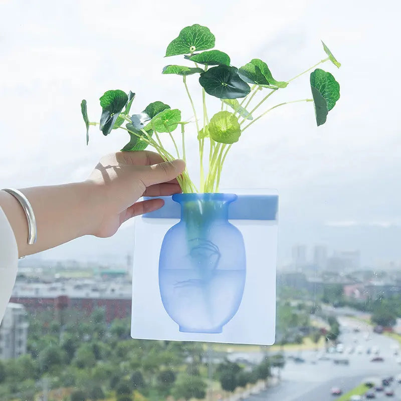 A hand holds a green plant in a blue self-adhesive wall flower vase against an outdoor blurred background.
