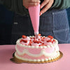 Una mano usa una boquilla de decoración de acero inoxidable rosa para decorar un pastel glaseado rosa con fresas.