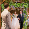 A bride and groom stand under a lush silk wisteria garland vine for a wedding ceremony.