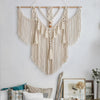 A cream-colored macrame wall hanging tapestry with fringe and tassels is displayed on a white wall.
