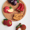 A set of realistic fruit and vegetable fridge magnets including a plum, apple, chili peppers, and a raspberry on a wooden boa