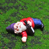 A miniature resin gnome statue with a white beard wearing a red shirt lying on green grass.