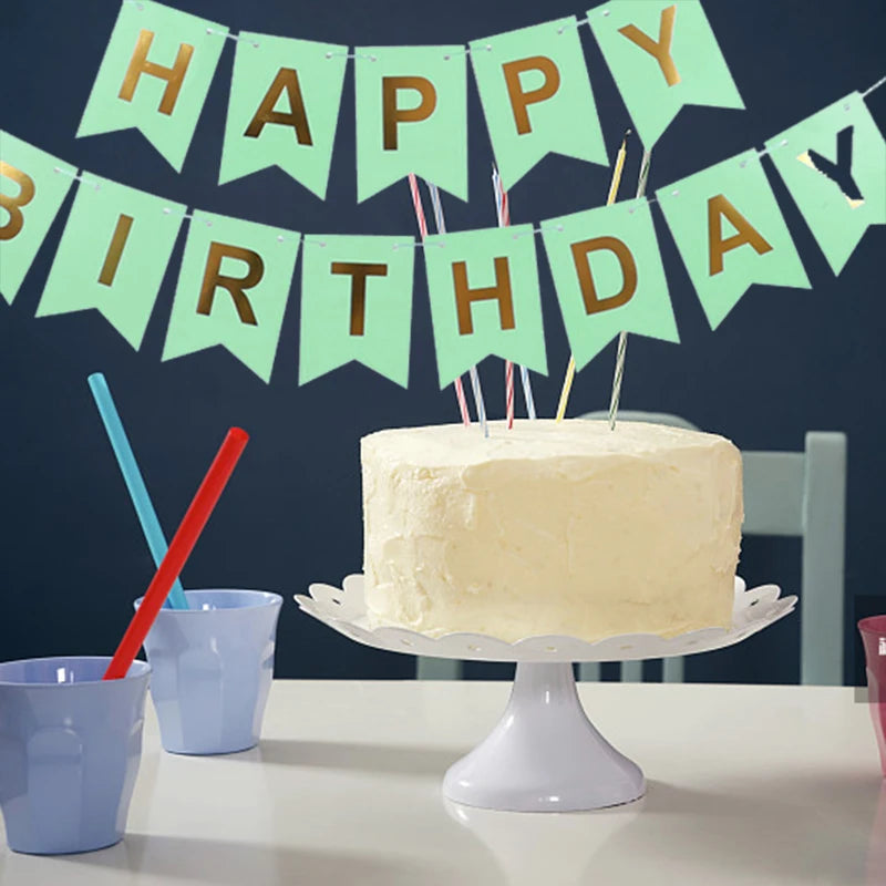 Un pastel de cumpleaños blanco decorado con banderas doradas y palitos de colores sobre una mesa.