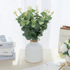 A bouquet of silk eucalyptus leaf stems in green and cream sits in a white vase on a light surface.