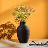A black ceramic vase filled with light yellow and pink flowers sits on a white surface against an orange wall.