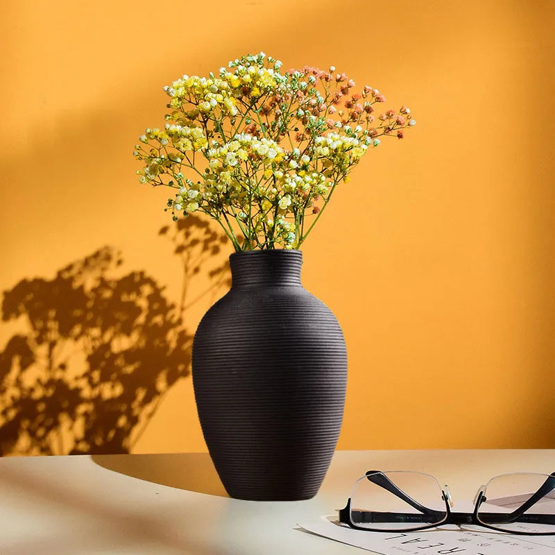 A black ceramic vase filled with light yellow and pink flowers sits on a white surface against an orange wall.