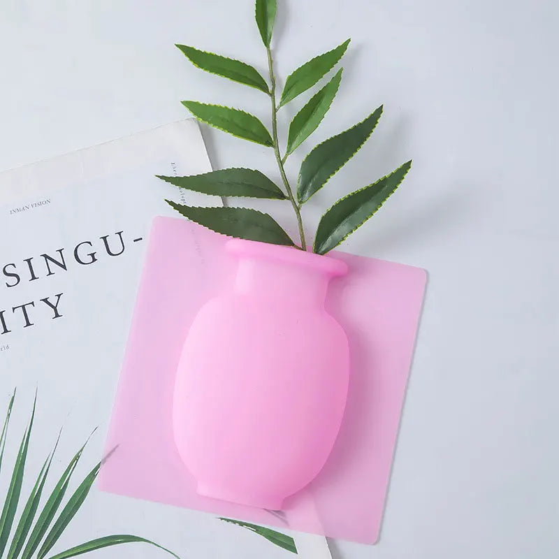 A pink flexible self-adhesive wall flower vase is shown resting on a pink square piece of material next to a green leafy bran