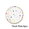A white disposable paper plate with a gold rim decorated with colorful cartoon party icons like balloons and cupcakes.