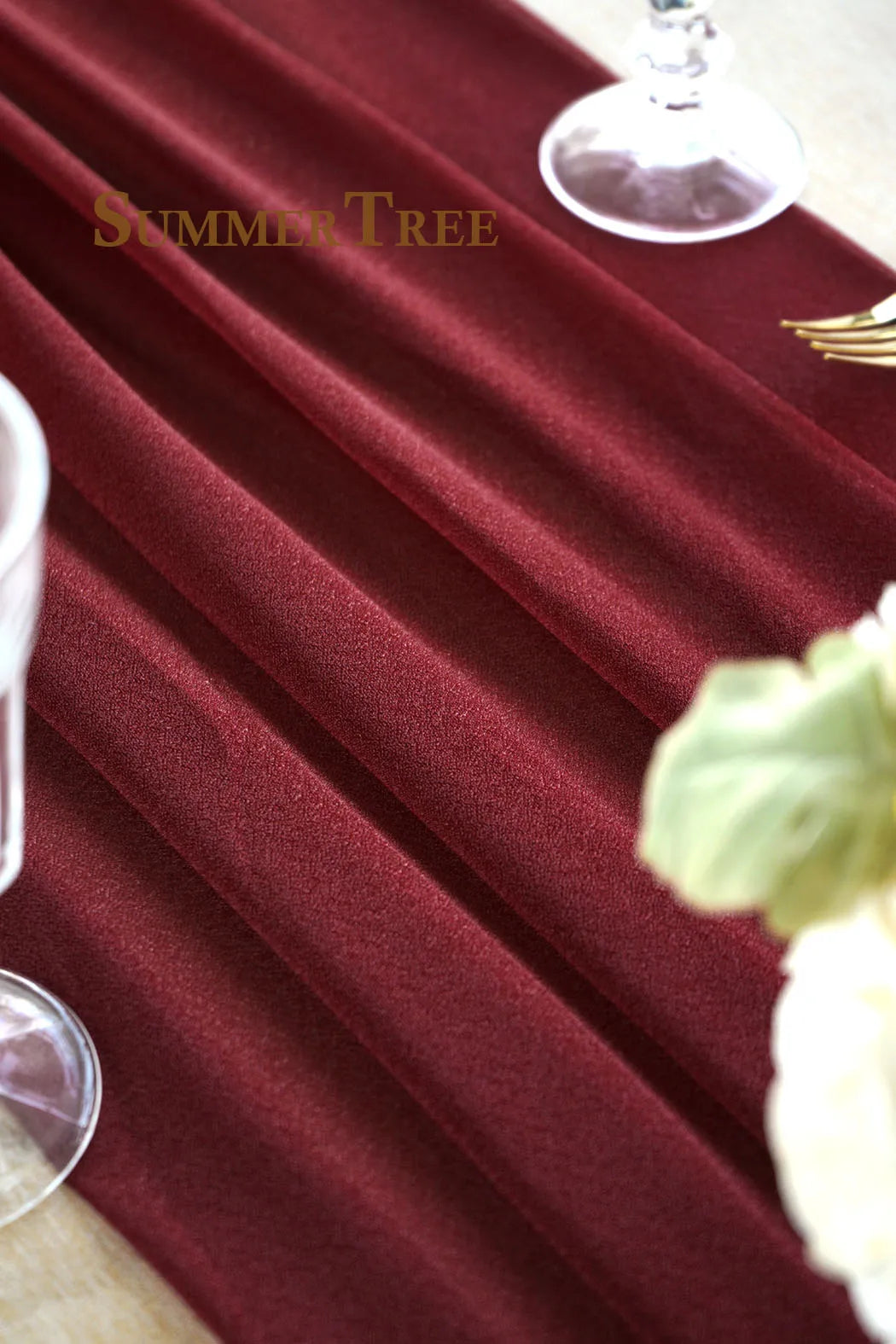 A close-up of deep burgundy chiffon fabric draped elegantly on a table, with a glass and a white flower visible.