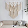 A beige macrame wall hanging tapestry with fringe details displayed on a white wall above a light-colored sofa.