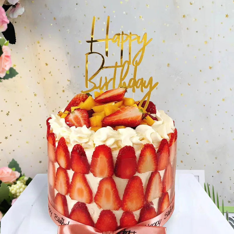 A round cake decorated with strawberries and cream, topped with gold acrylic happy birthday toppers.