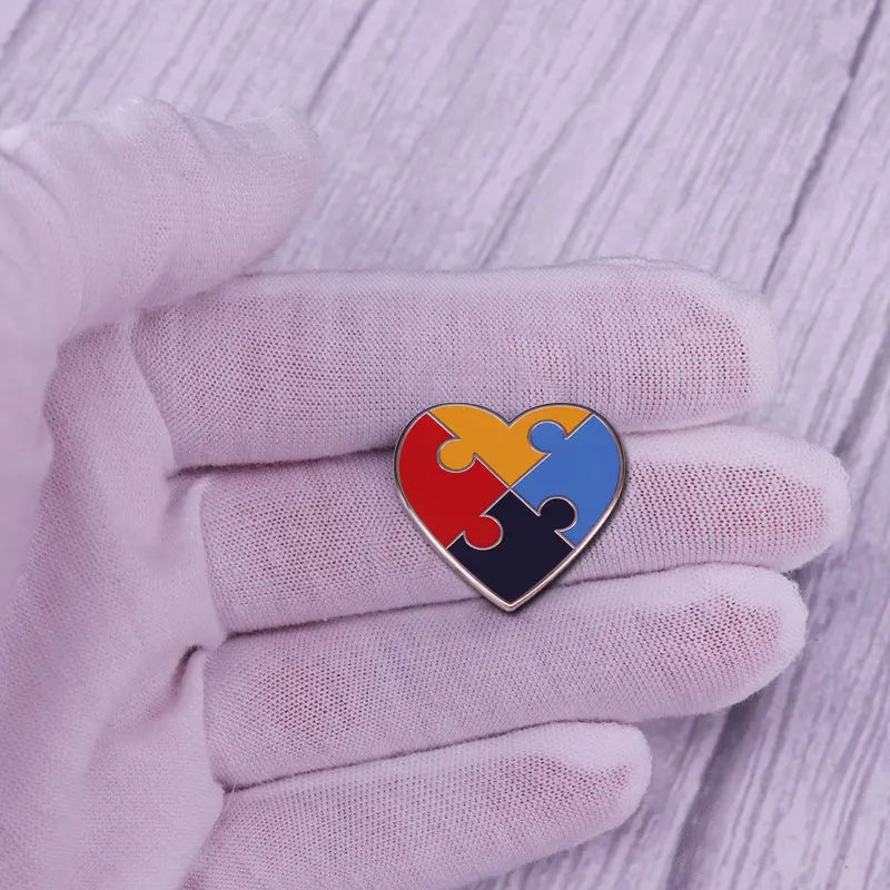 A hand wearing a light purple glove holds a heart-shaped puzzle ribbon brooch pin with rainbow colors.