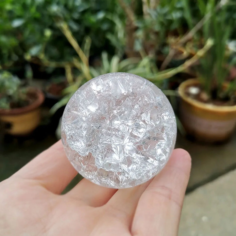 A hand holds a clear, spherical crystal ball water feature ornament against a blurred green plant background.