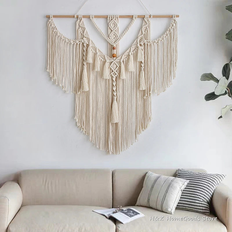 A beige macrame wall hanging tapestry with fringe and tassels is displayed on a white wall above a light-colored sofa.