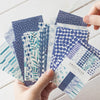 A hand holds a stack of decorative stationery paper featuring various patterns and shades of blue and teal.