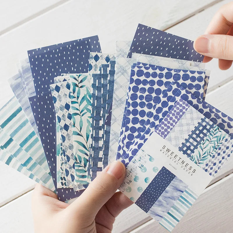 A hand holds a stack of decorative stationery paper featuring various patterns and shades of blue and teal.
