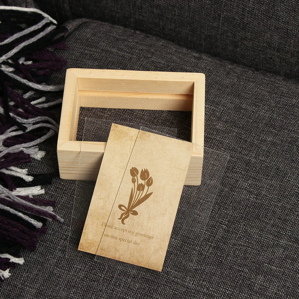 A light wooden photo frame holds a vintage card with a floral design and text on a dark gray fabric.