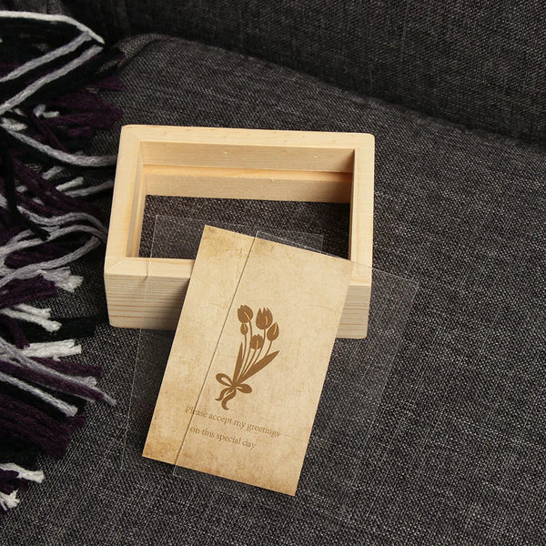 A light wooden photo frame holds a vintage card with a floral design and text on a dark gray fabric.
