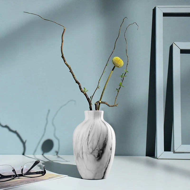 A white marble ceramic vase holds delicate, bare branches with a single yellow flower against a light blue wall.