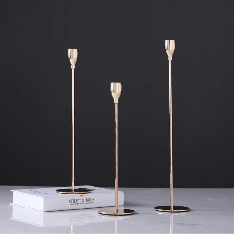 A set of three slender gold-toned iron candle holders displayed on a white book against a dark background.