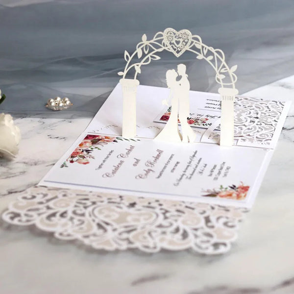 Invitaciones de boda de perlas doradas cortadas con láser, con un arco blanco y silueta de una pareja sobre una superficie de