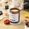 A jar of marigold scented candle sits on a light gray coaster next to dried flower petals and a glass bottle.