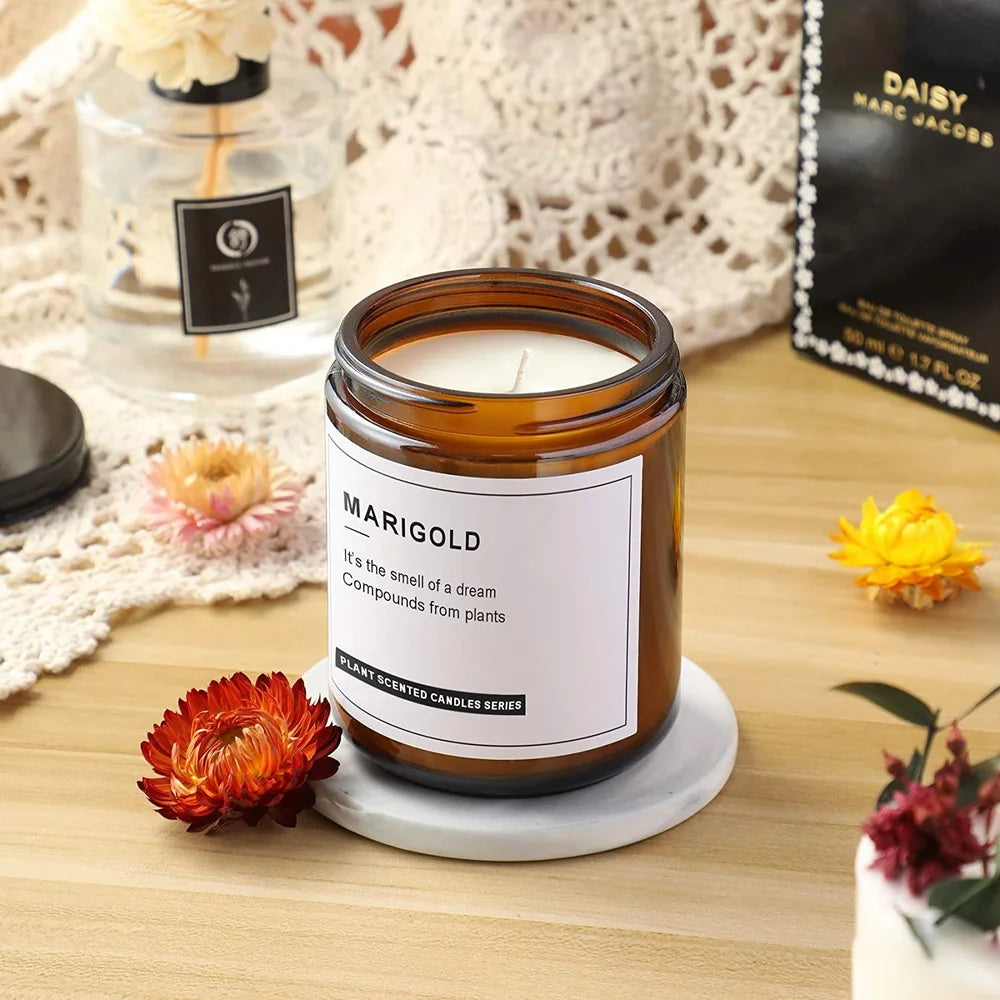 A jar of marigold scented candle sits on a light gray coaster next to dried flower petals and a glass bottle.