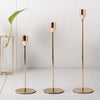 Three slender gold-toned iron candle holders standing on a white surface.