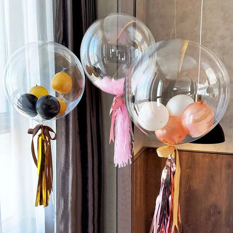 A bundle of clear balloons in various shades of pink and gold are displayed against a dark curtain.