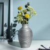 A gray striped ceramic vase holds yellow and white flowers against a light blue wall.