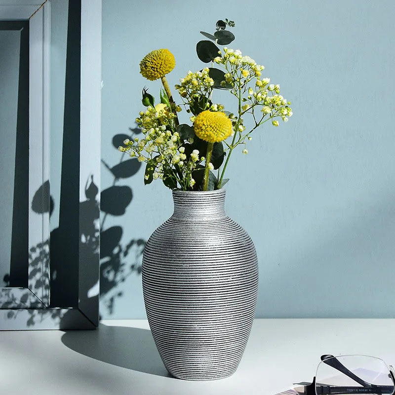 A gray striped ceramic vase holds yellow and white flowers against a light blue wall.