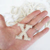 A hand holds a sparkling rhinestone letter X patch against a background of other pearl letters.