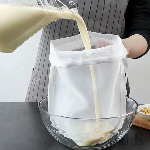A hand pours a light-colored liquid from a jug into a white reusable fine mesh nylon filter bag placed over a glass bowl.
