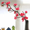 Pink artificial silk cherry blossom branches with vibrant red flowers displayed in a vase against a bright background.